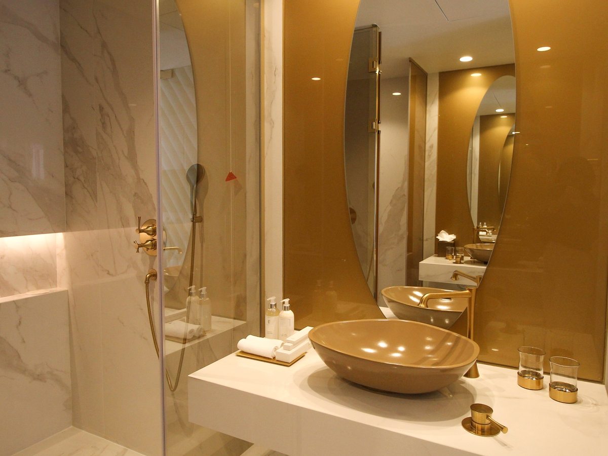 A marble bathroom with a glass walk-in shower, a stone basin on a white counter and an oval mirror set against a gold panel.