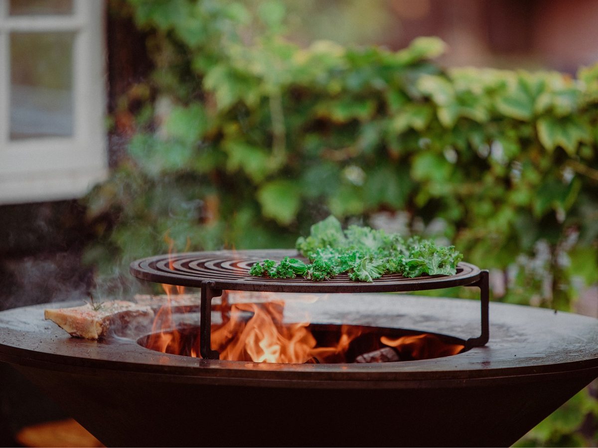 A round steel fire bowl with a grill plate of meat and herbs smoking over an open flame, on a garden terrace backed by greenery.