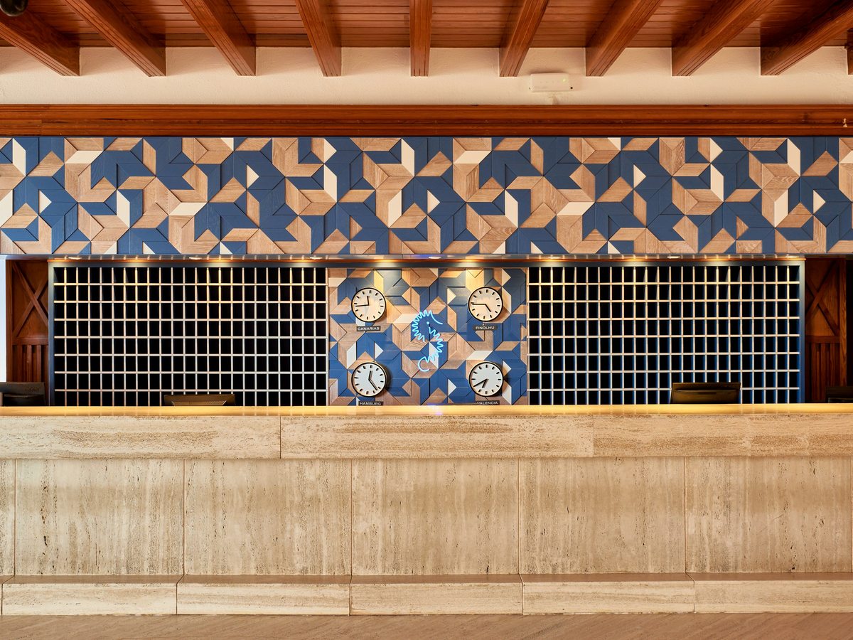 Long travertine reception counter below a blue and sand geometric tiled wall set with a row of world clocks.