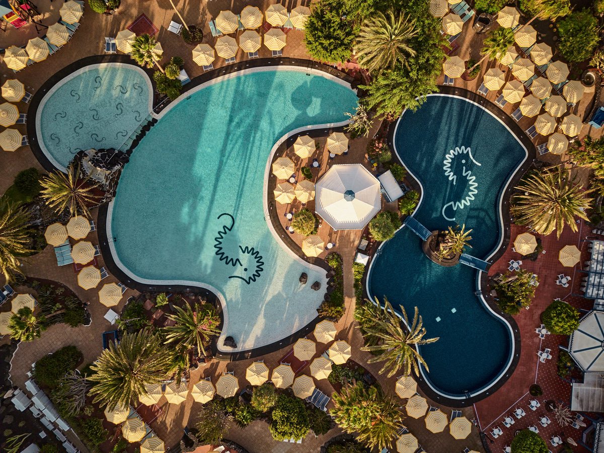 Aerial view of two curved pools with seahorse motifs on their floors, ringed by yellow parasols, sun loungers and palms.