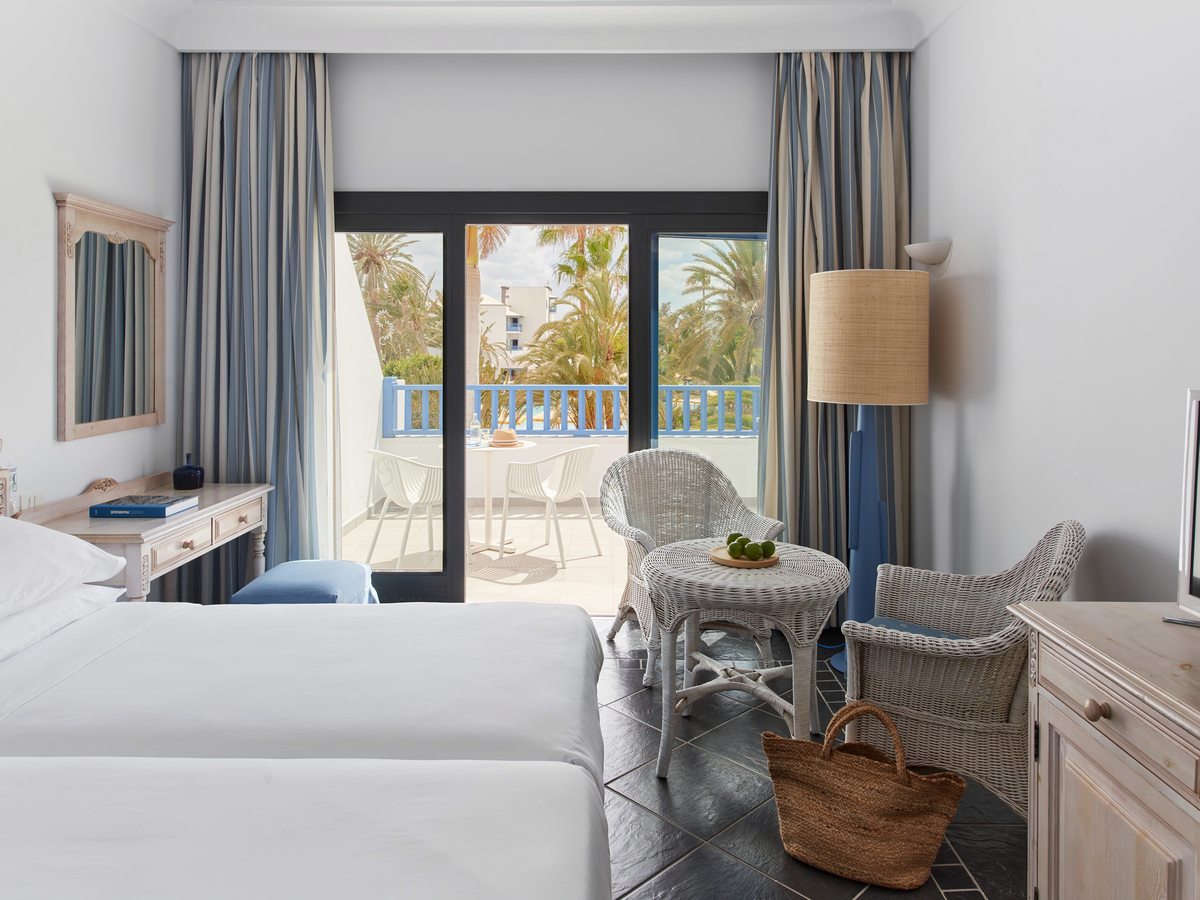 Twin room with the two beds pushed together, a pale wooden dresser and glass doors onto a balcony with wicker chairs.