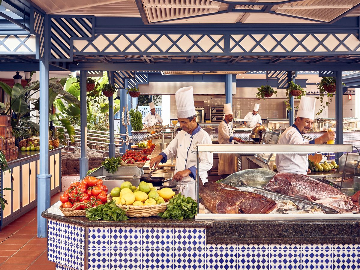 Chefs working at an outdoor buffet counter of fresh fish, tomatoes and lemons under a blue and white timber canopy.
