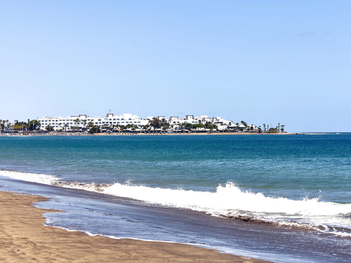 Wide sandy beach curving round a bay with breaking waves and low white hotel buildings along the shore.