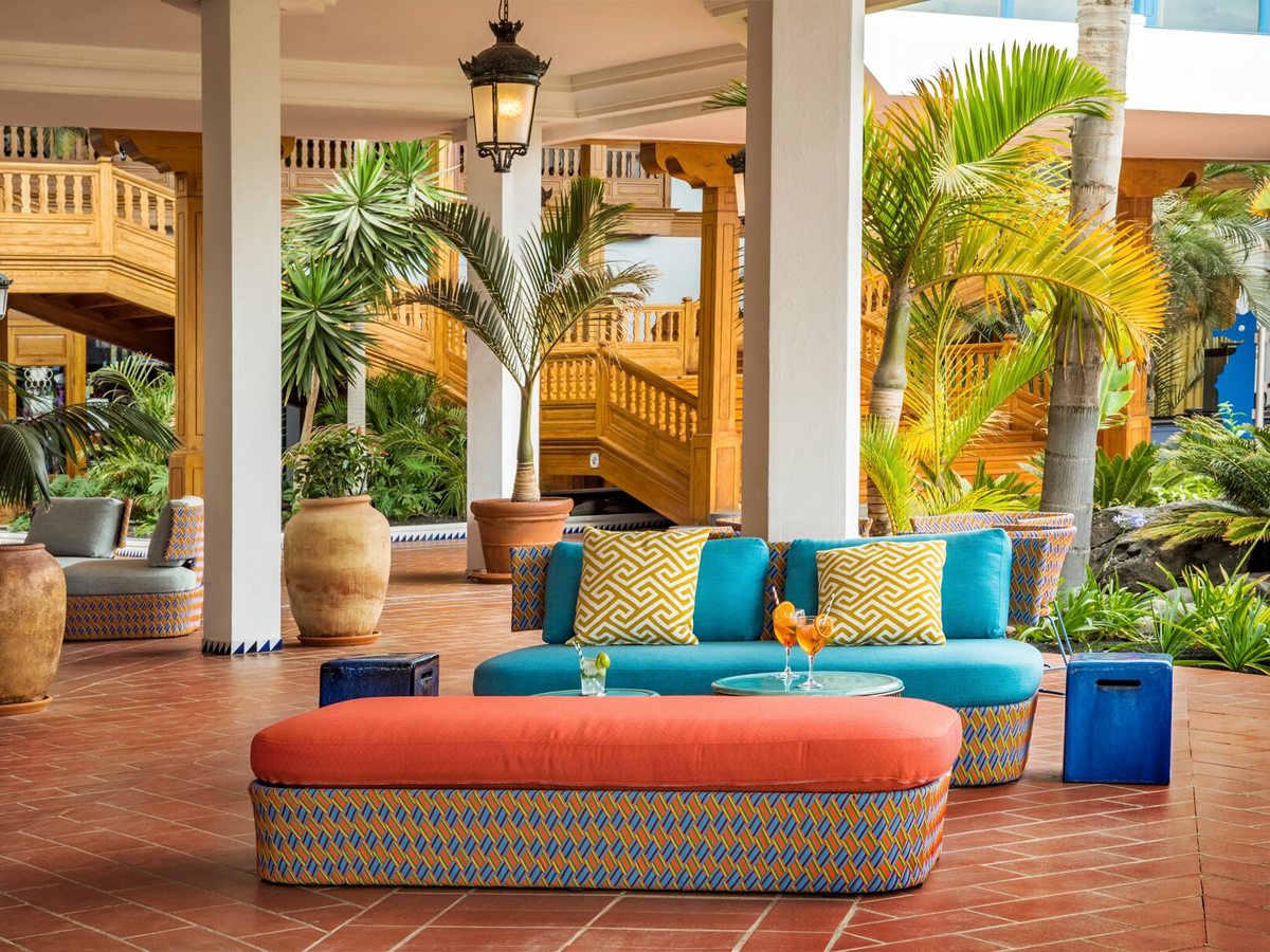 Open-air terrace with turquoise and coral cushioned seating and low tables among potted palms, beneath wooden balconies and hanging lanterns.