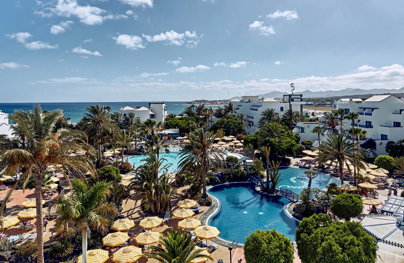The pools and palm gardens at Seaside Los Jameos, with low white buildings and the sea at Los Pocillos beyond.