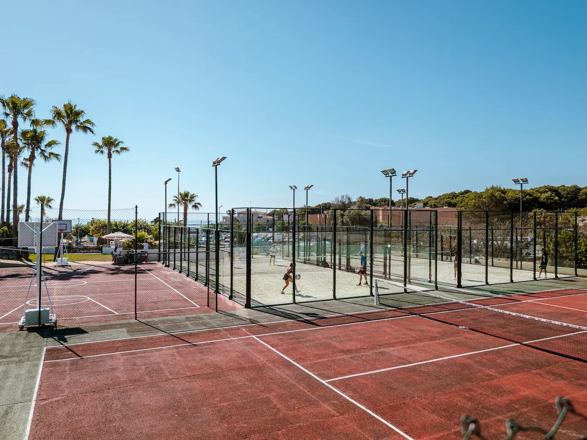 Fenced paddle tennis courts with players inside, a red hard court with a basketball hoop alongside and floodlight columns and pines behind.