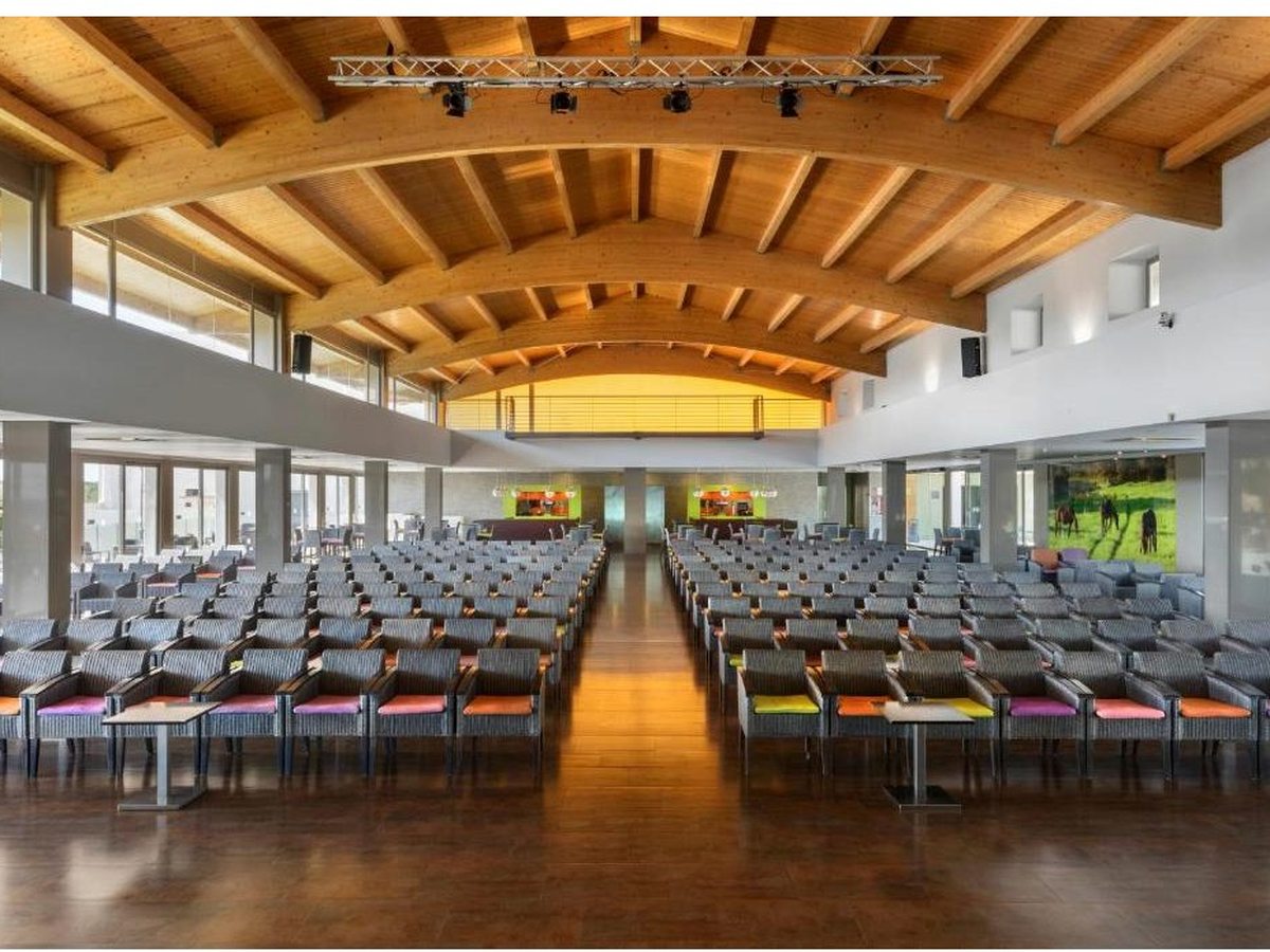 Rows of grey and orange seats facing a stage under a curved timber-beamed ceiling hung with lighting rigs.