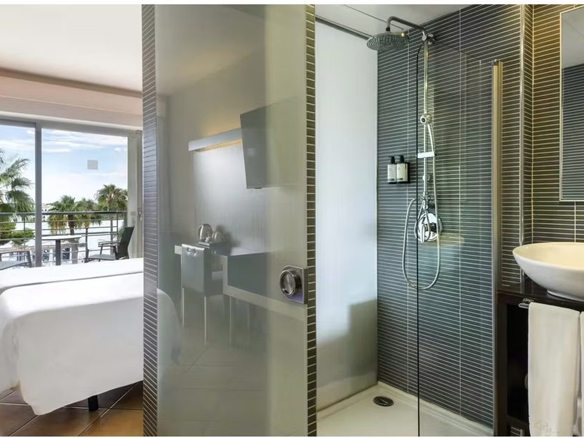 A double room with a white-linen bed, an open glass shower and washbasin beside it, and balcony doors onto a terrace with palms.