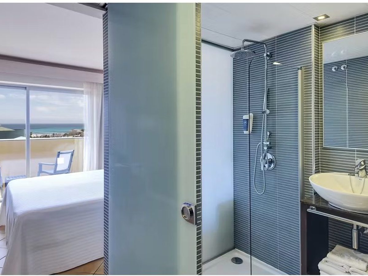A double room with the sea through open balcony doors, and an open glass shower and washbasin alongside the bed.