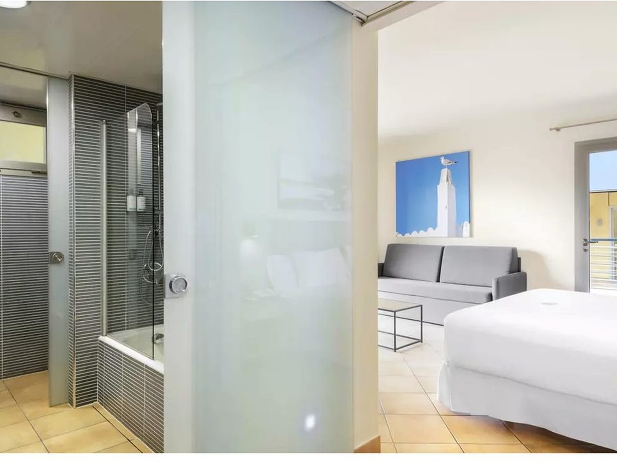 A deluxe room with a bed, a grey sofa and a low table, and a bathroom with a shower over the bath behind a glass screen.