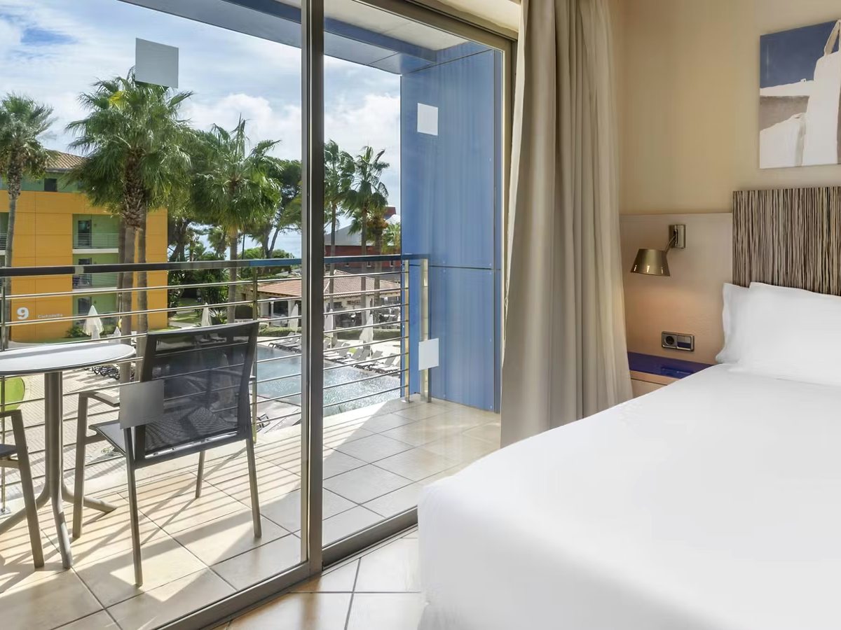 A family room bedroom with balcony doors open onto a table and chairs, with palms and a pool below.