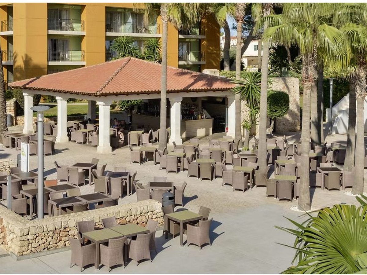 A tile-roofed open bar pavilion with white columns, ringed by wicker chairs and tables on a paved terrace among palms.