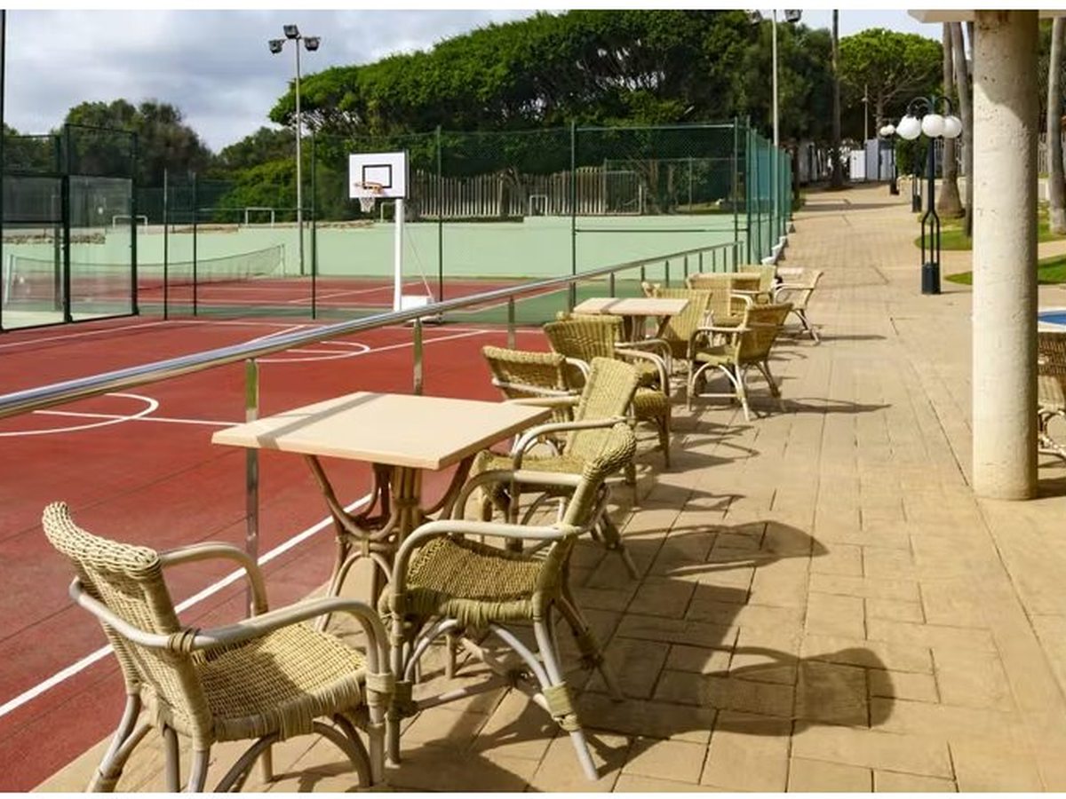A red multi-sport court with basketball hoops behind green fencing, with wicker chairs and tables along the paved walkway beside it.