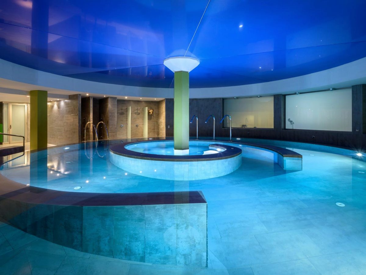 An indoor spa pool with a circular whirlpool at its centre under a domed blue-lit ceiling, with steps and handrails at the edge.
