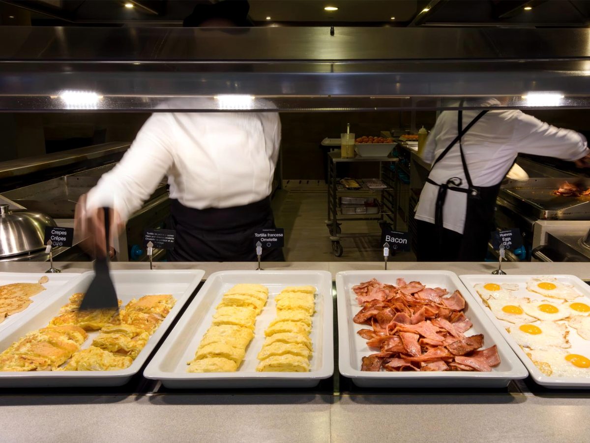 Two chefs in white jackets working behind a service counter set with trays of omelette, bacon and fried eggs.