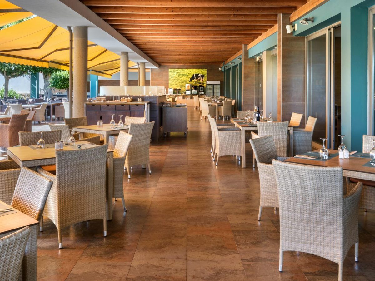 Buffet restaurant dining room with cream wicker chairs at wooden tables on a tiled floor, and a yellow awning shading the terrace doors.