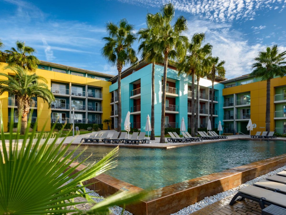 A turquoise pool with a raised spillway edge in front of it, sun loungers along the far side and palms in front of the colourful blocks.