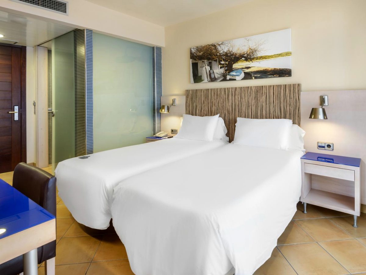 Twin beds side by side under a woven headboard, with bedside tables, wall lamps and a framed landscape picture above.