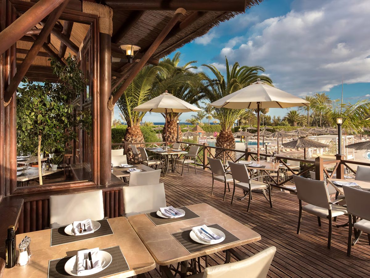 A raised wooden restaurant terrace under a thatched roof with tables laid for lunch, looking across parasols and the pool to the sea.