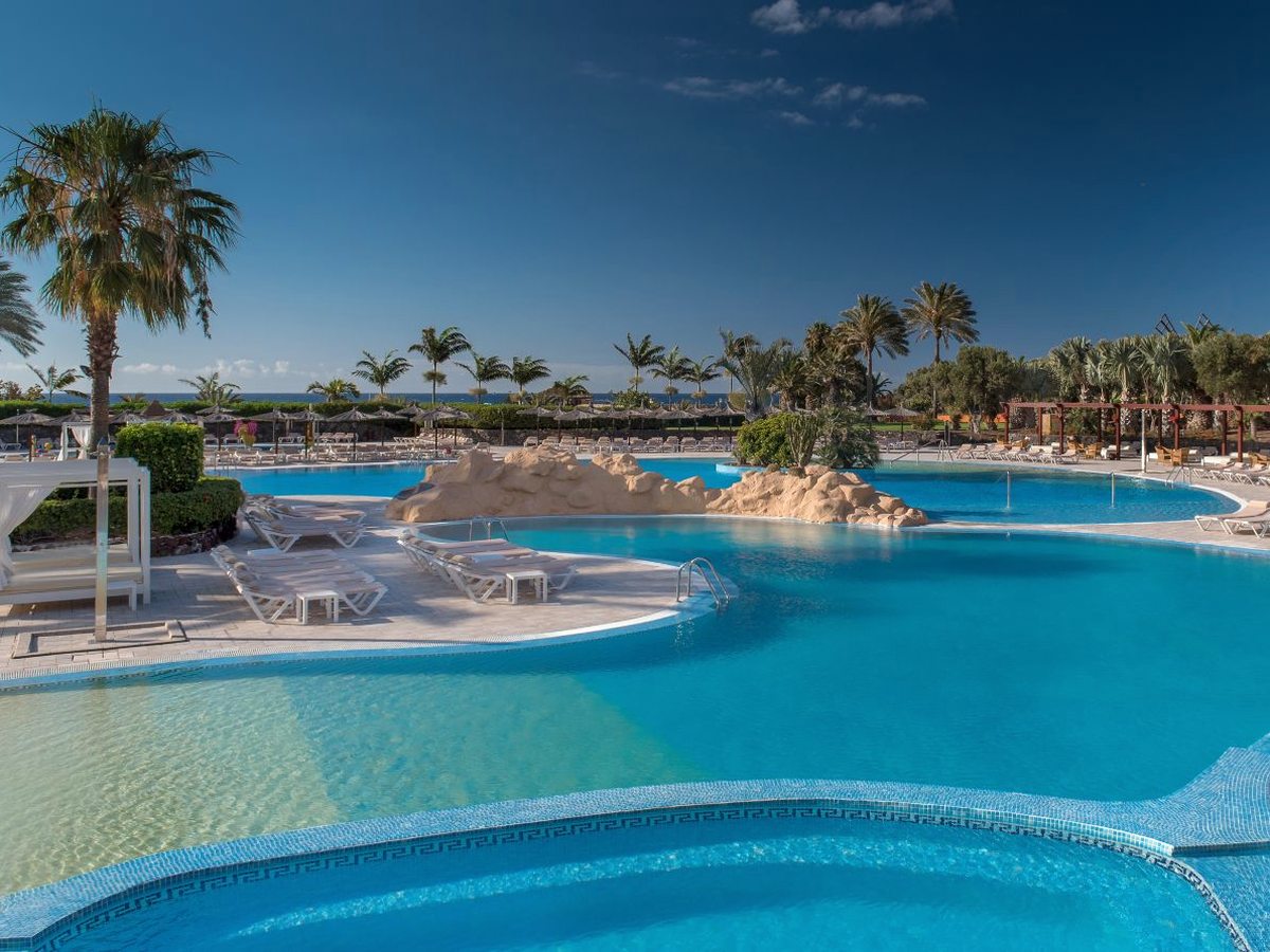 A pool with rock features and a curtained cabana, loungers along its edge and rows of thatched parasols beyond, with the sea on the horizon.