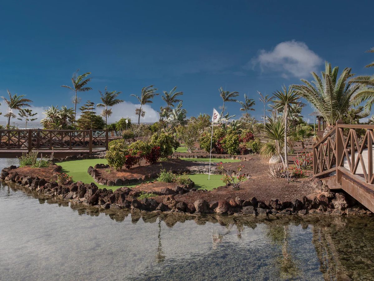 A mini-golf hole with an artificial green and a numbered flag on a volcanic-rock island, a wooden footbridge crossing the shallow water in front of it and palms behind.