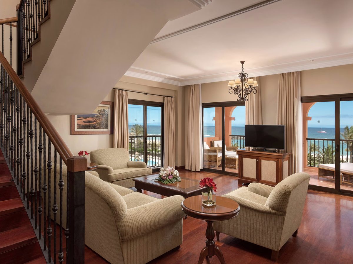 A suite lounge with armchairs around a coffee table, an internal staircase with a wrought-iron balustrade, and balcony doors onto the sea.