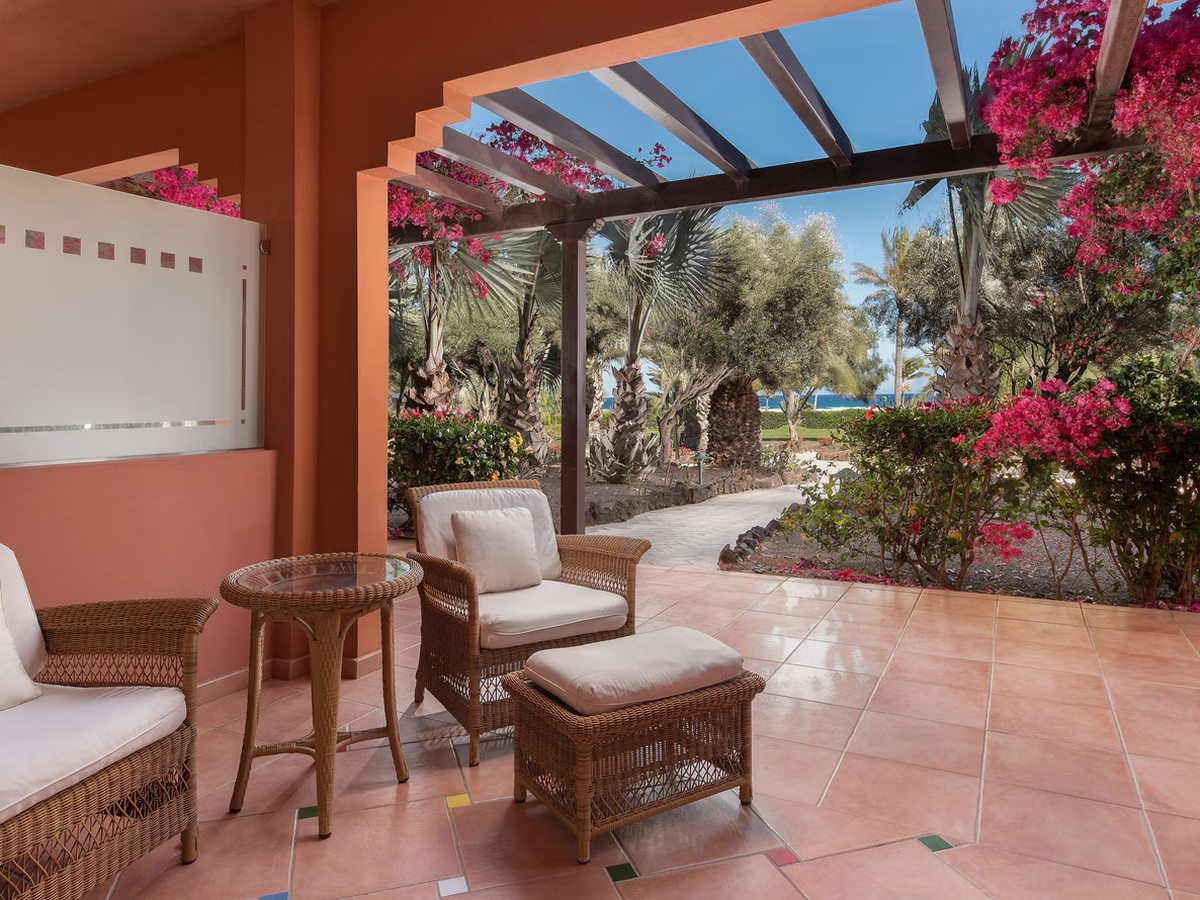 A ground-floor terrace with wicker chairs and a small table under a timber pergola hung with bougainvillea, opening onto a palm garden.