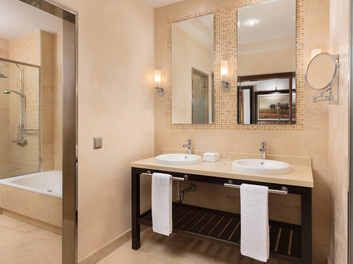 A bathroom with twin basins set in a stone-topped vanity below a large mosaic-framed mirror, and a bath with a glass shower screen through the doorway.
