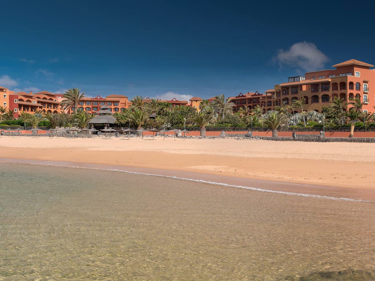 A wide golden-sand beach with clear shallow water in the foreground, palms and thatched parasols along the top of the sand and the resort behind.