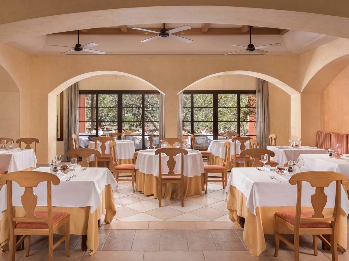 An arched indoor dining room with white-clothed round and square tables, wooden chairs, ceiling fans and tall windows onto a garden terrace.