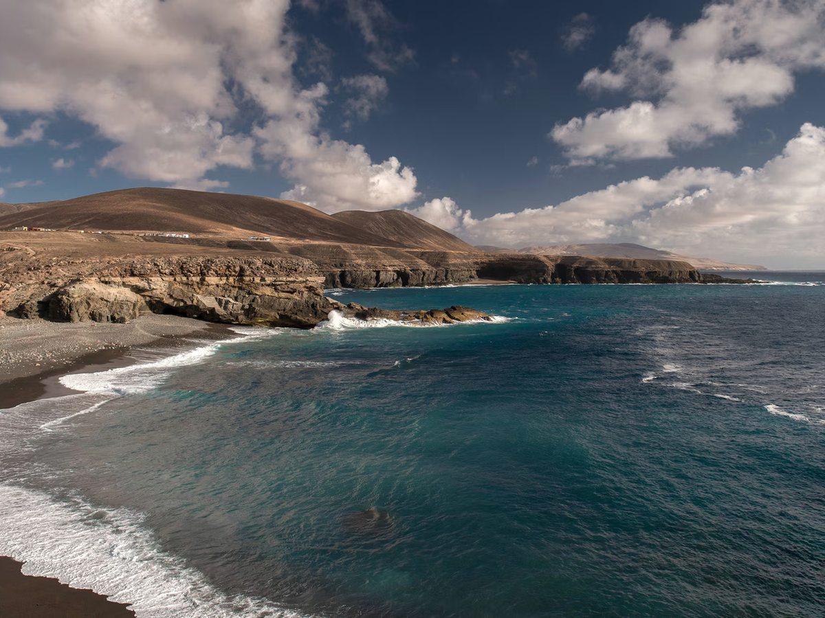 A dark-sand beach below low rock cliffs, with waves breaking over rocks and brown hills running away along the coast.