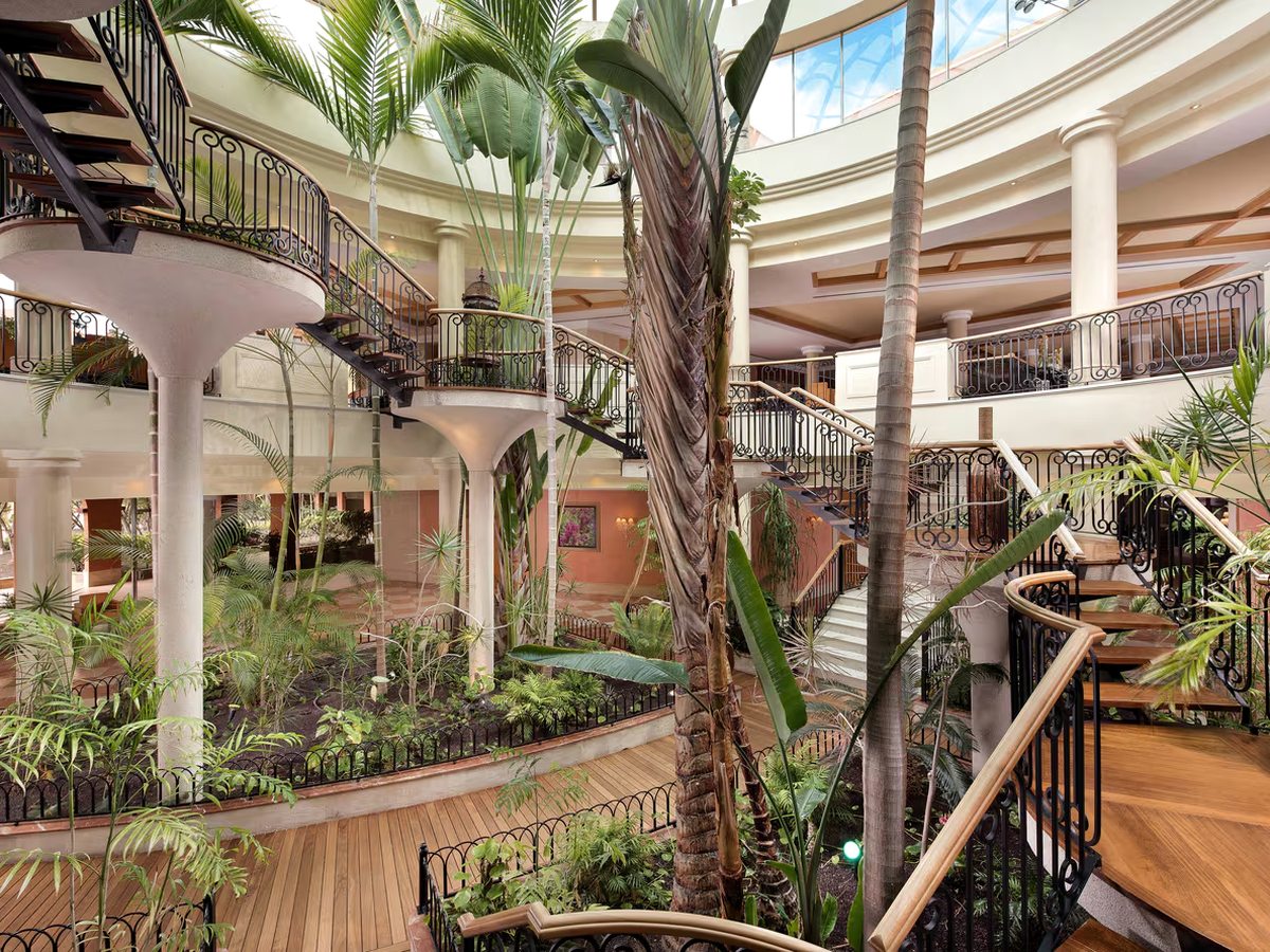 A multi-level indoor atrium planted with tall palms and ferns, with curved staircases and wrought-iron balconies, lit from a glazed roof above.
