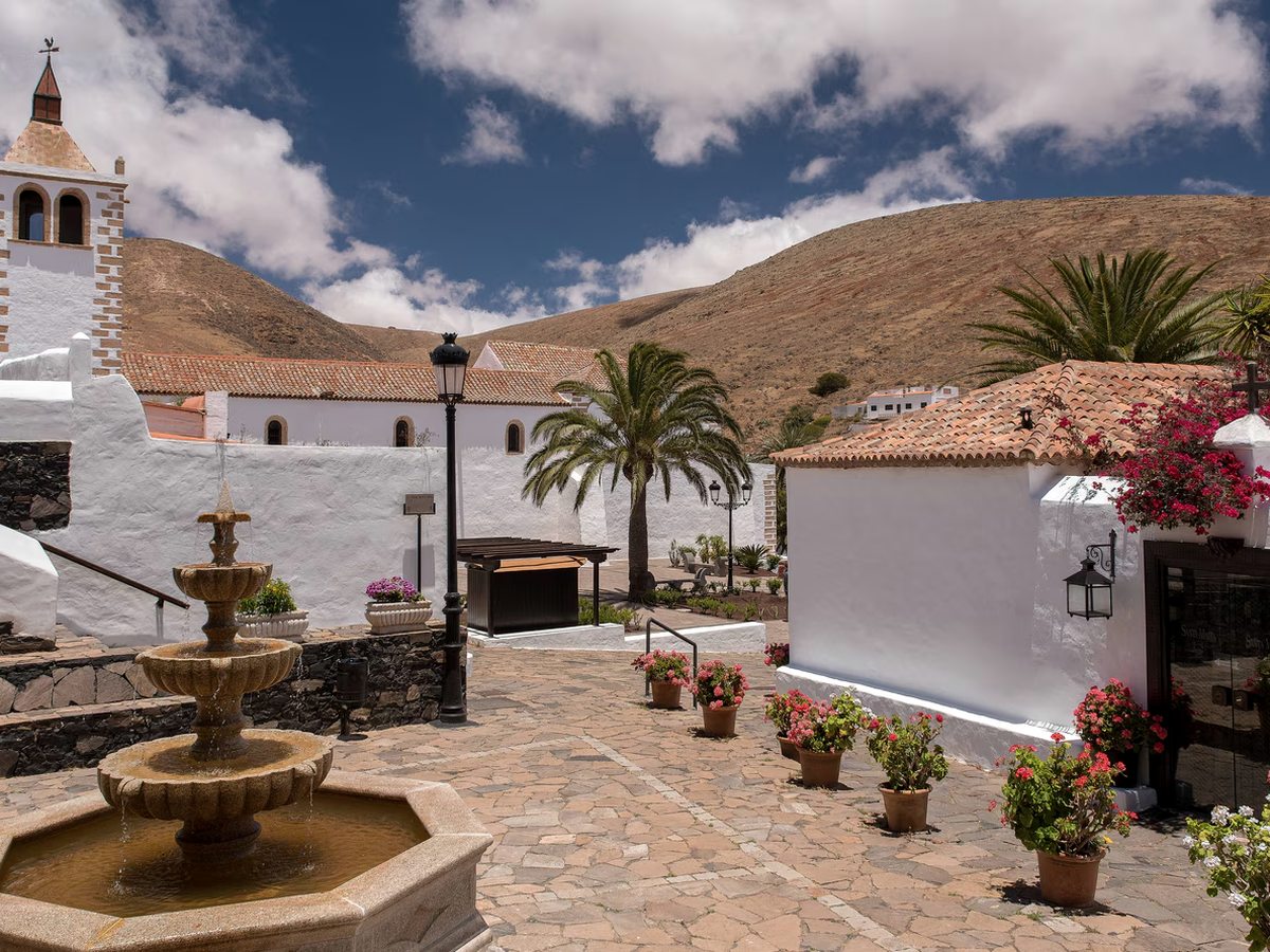 A cobbled village square with a stone fountain, a whitewashed church with a bell tower, potted geraniums and bare brown hills behind.