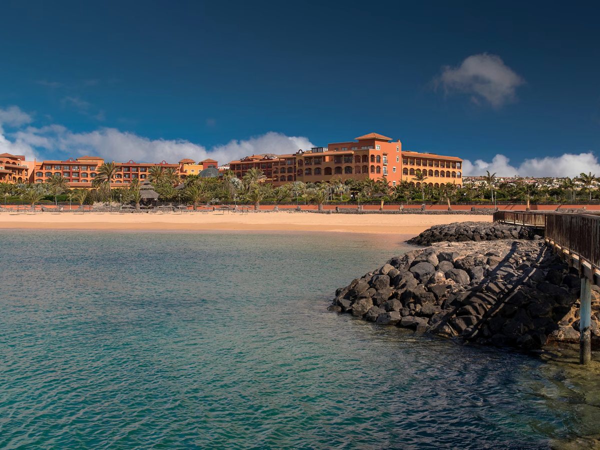 A dark rock breakwater and wooden jetty at the end of a sandy bay, with calm turquoise water and the resort buildings along the shore.