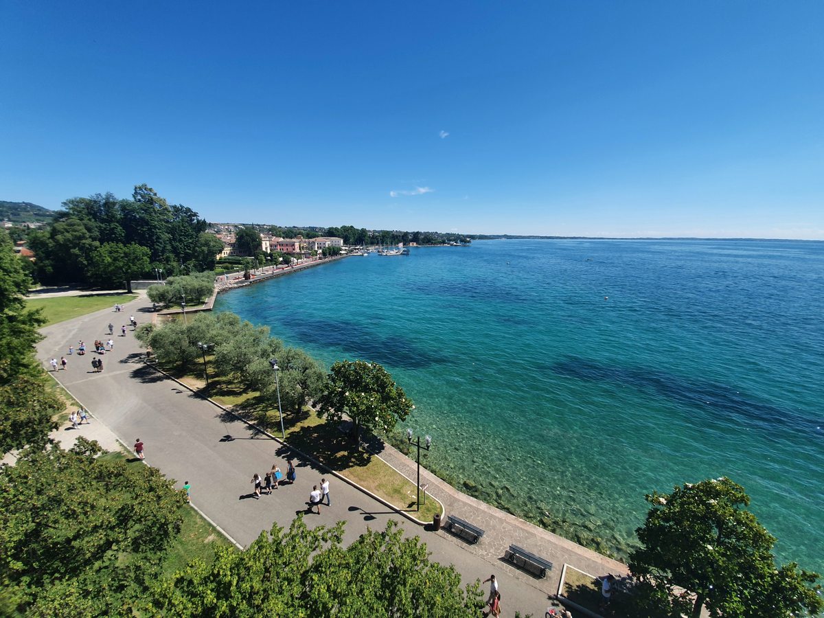 A wide view along the shore of Lake Garda: a tree-lined promenade with benches curving towards a small town and its marina, and clear turquoise water shading to deep blue.