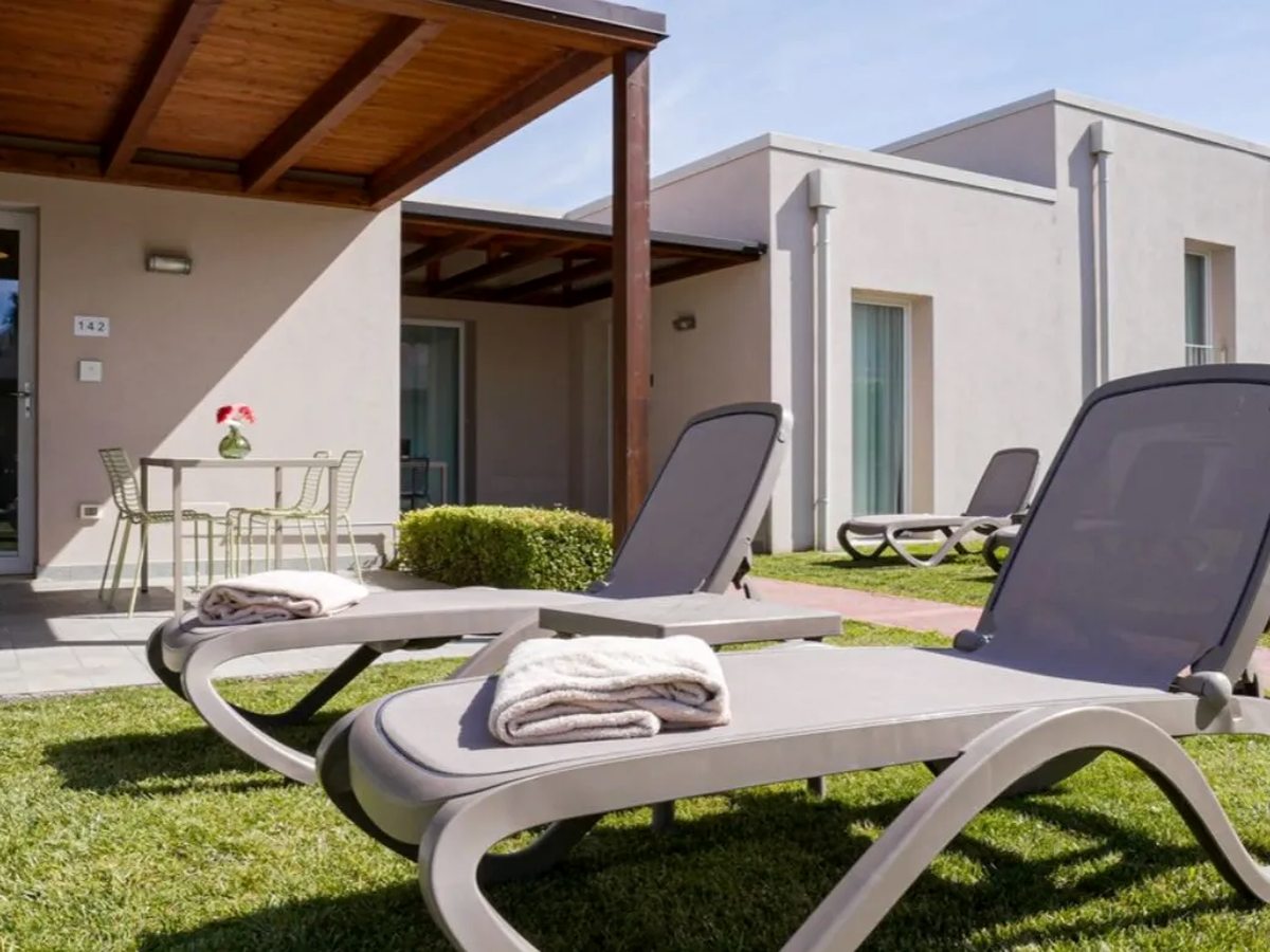 A single-storey apartment with a timber-framed covered patio, a small table and pale green chairs by the door, and two grey sun loungers with folded white towels on the lawn in front.