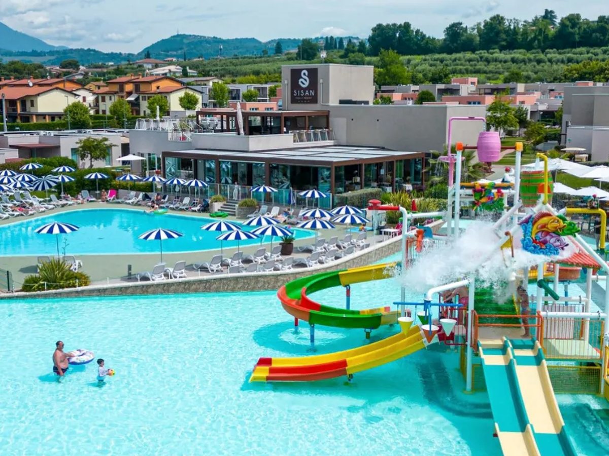 A wide turquoise lagoon pool with a brightly coloured water-play structure and green, red and yellow slides, an adult and a small child standing in the shallow water, and the Sisan restaurant building and its rooftop sign behind.