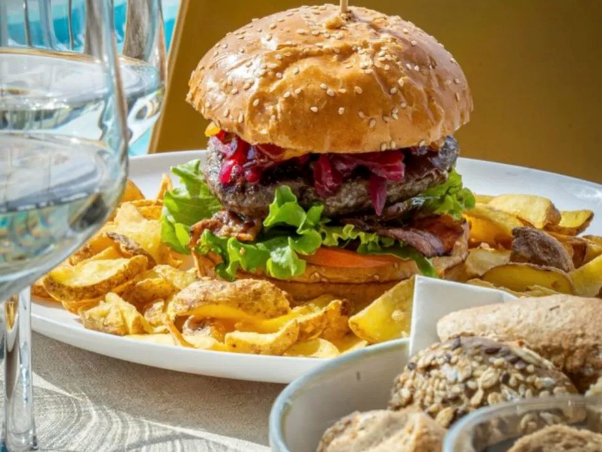 A beef burger in a sesame bun with lettuce, tomato and red onion, served on a white plate with a pile of golden potato crisps, a basket of bread rolls alongside and a wine glass in front.