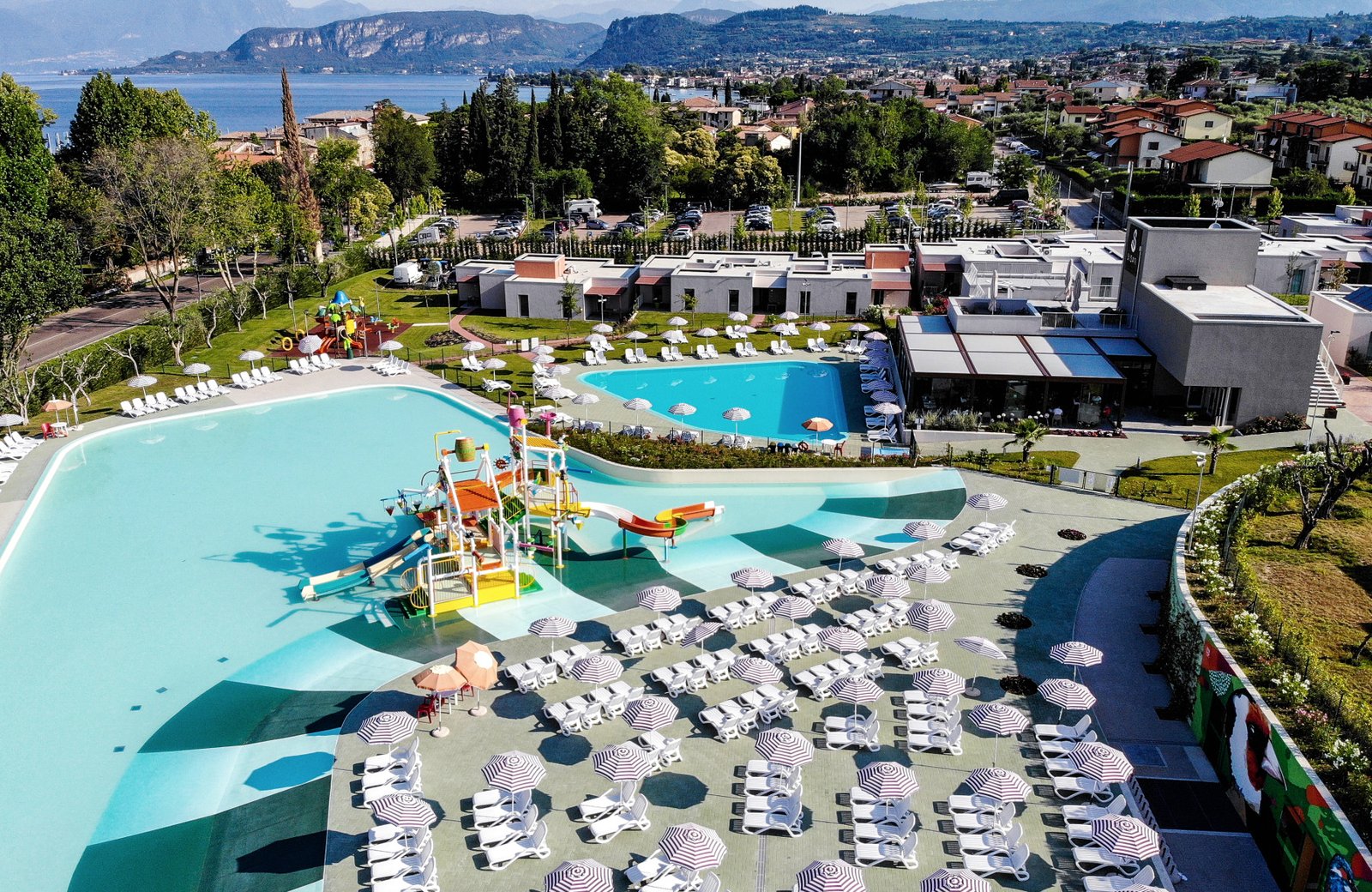 Aerial view of Sisan Family Resort at Cisano di Bardolino: the turquoise lagoon pool and its water park, a second swimming pool beside the restaurant, white apartment blocks in gardens, and Lake Garda and the mountains beyond.