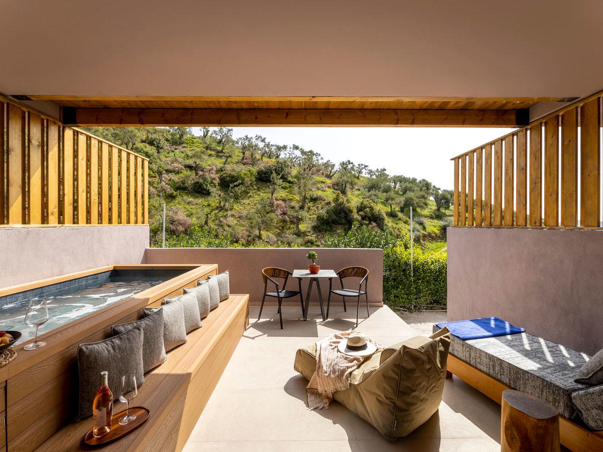 A covered terrace with a long built-in jacuzzi, a timber bench lined with cushions, bean bags, a cushioned daybed and a small dining table, with a green hillside beyond.