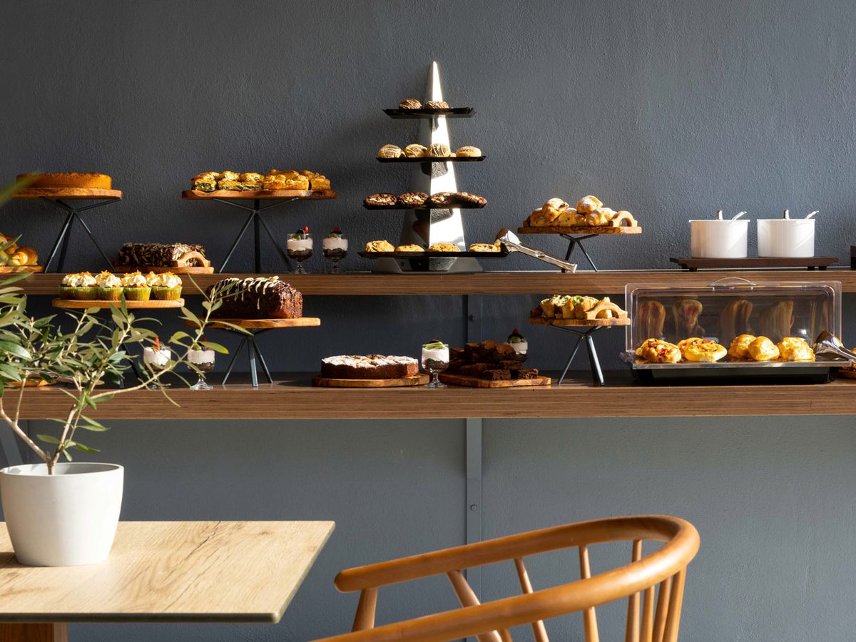 A long wooden buffet shelf against a grey wall holding cakes, tarts, savoury pastries, muffins and small desserts on wooden boards and stands.
