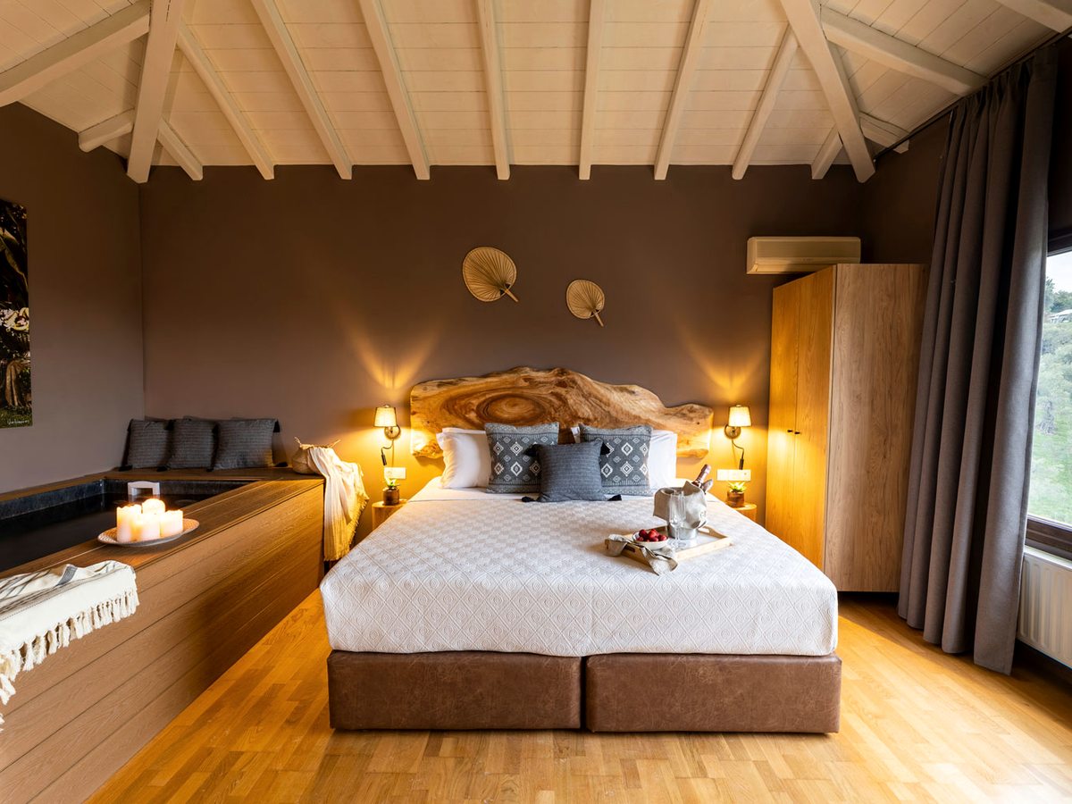 A bedroom under a white-beamed pitched ceiling, with a live-edge wooden headboard, wall lamps and a sunken jacuzzi set into a timber surround along one side, candles on its edge.