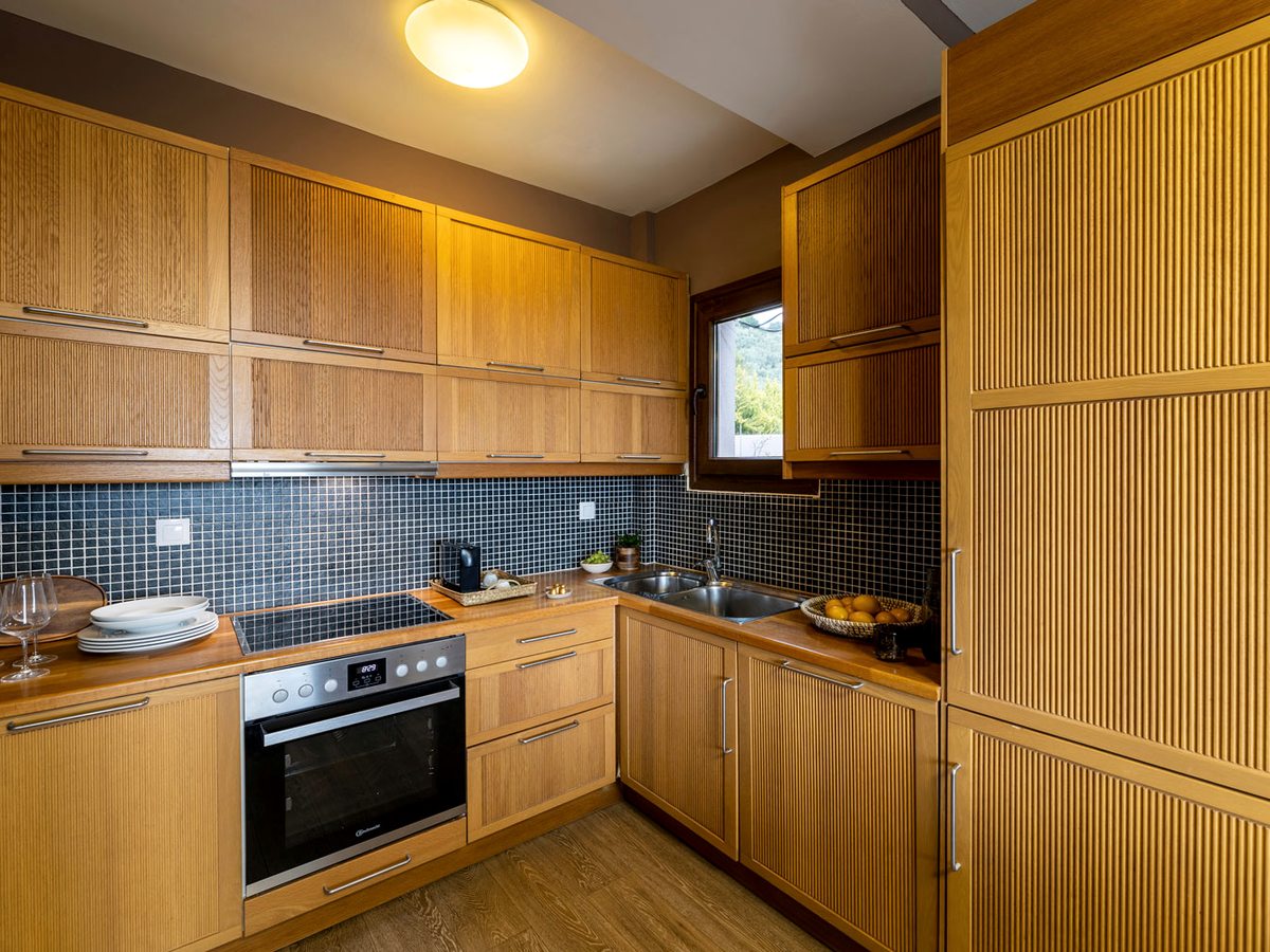 A fitted kitchen with fluted wooden units, a dark mosaic-tiled splashback, an oven, hob and sink, with plates and a bowl of oranges on the worktop.
