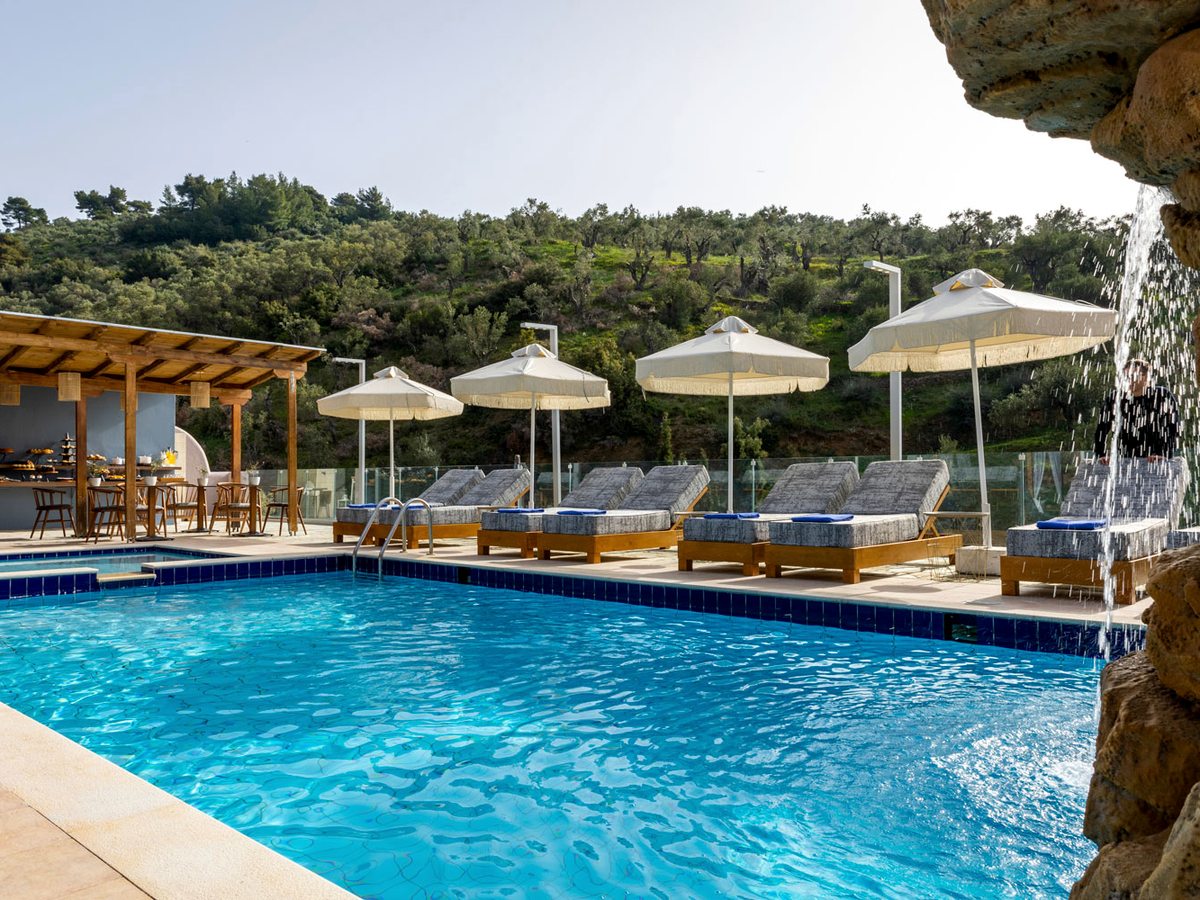 A large outdoor pool with a rock waterfall at one end, a row of grey sun loungers with blue towels under fringed white parasols, an olive-covered hillside behind and a covered bar to the left.
