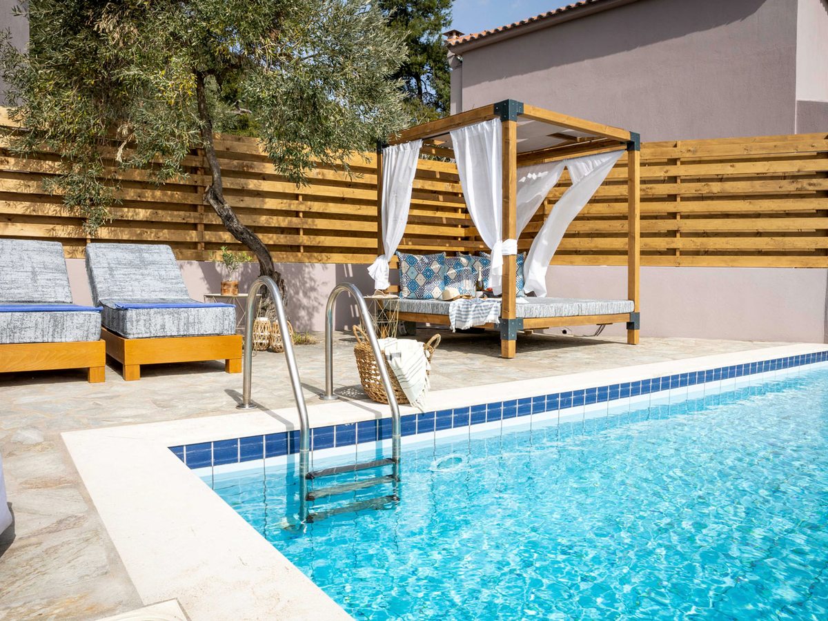 A small rectangular pool with a metal ladder, edged by a paved terrace with sun loungers, a curtained daybed and a slatted wooden privacy screen, with an olive tree behind.