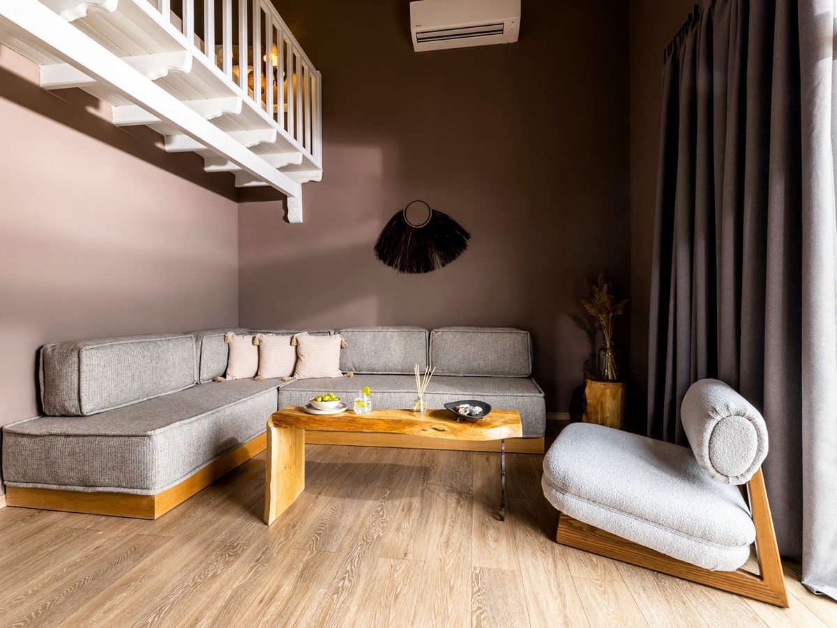 A grey corner sofa and a live-edge wooden coffee table in a lounge with a boucle armchair, a white staircase rising to a mezzanine and full-length curtains.