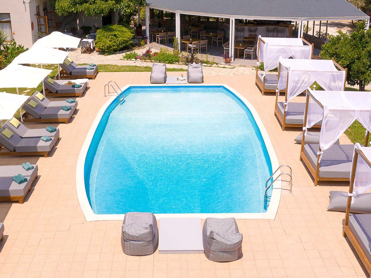 Aerial view of an outdoor pool with sun loungers under parasols along one side, curtained wooden daybeds along the other and a canopied dining terrace behind.
