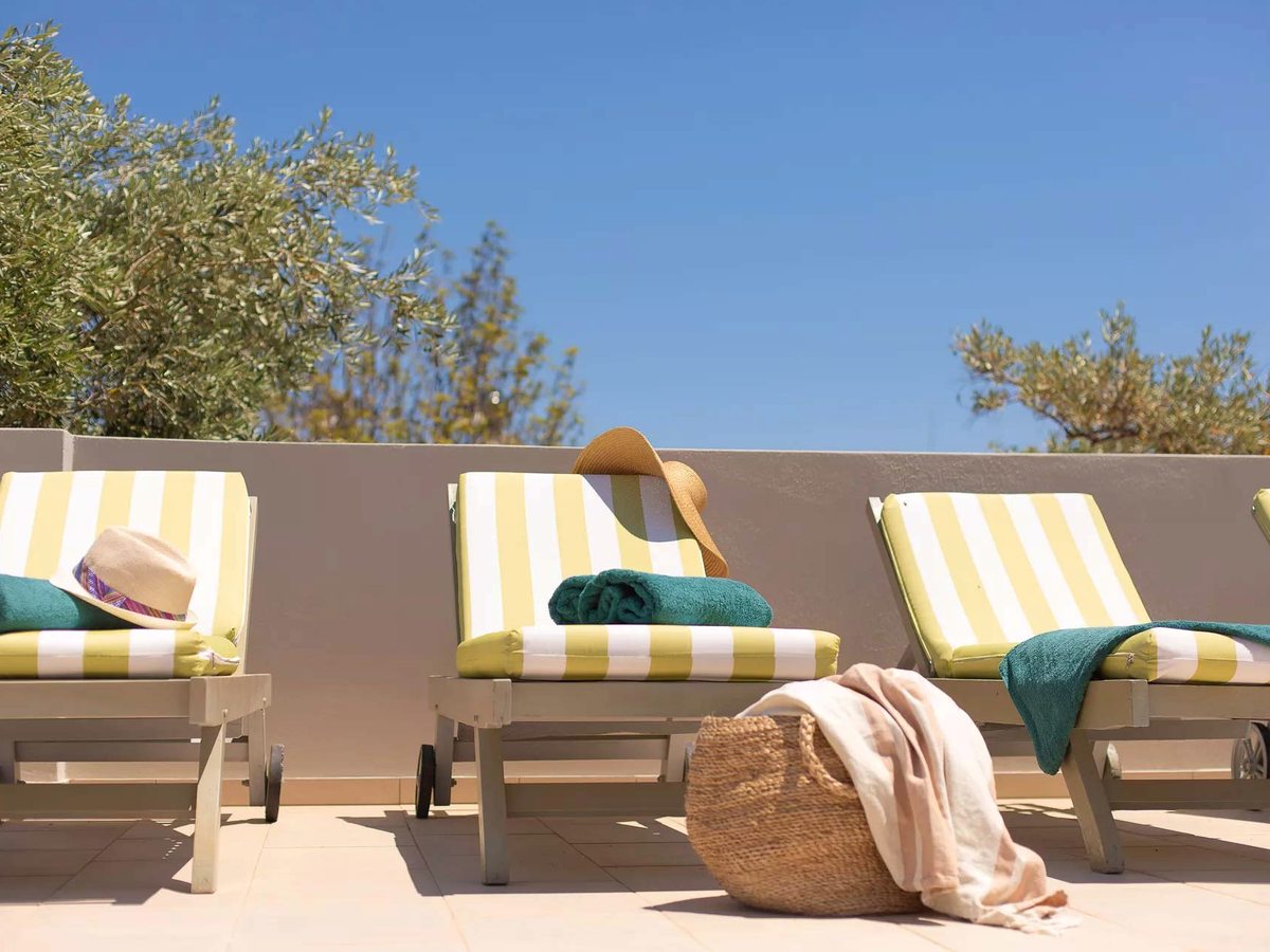 A row of wooden sun loungers with yellow and white striped cushions on a paved terrace, with towels, a straw hat and a woven basket beside them under a clear blue sky.