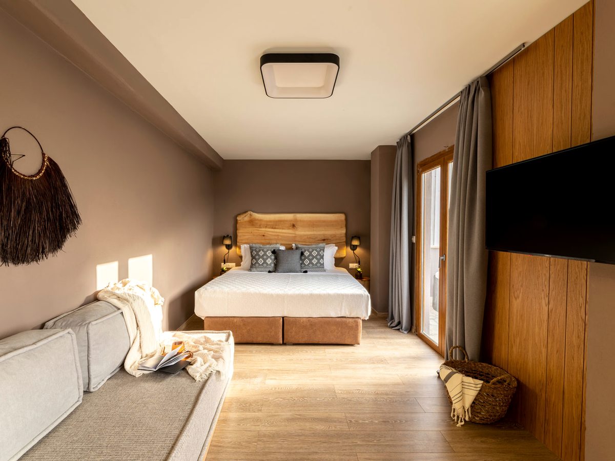 A suite with a double bed against a live-edge wooden headboard, a long sofa along the opposite wall, a wall-mounted television on timber panelling and curtained balcony doors.