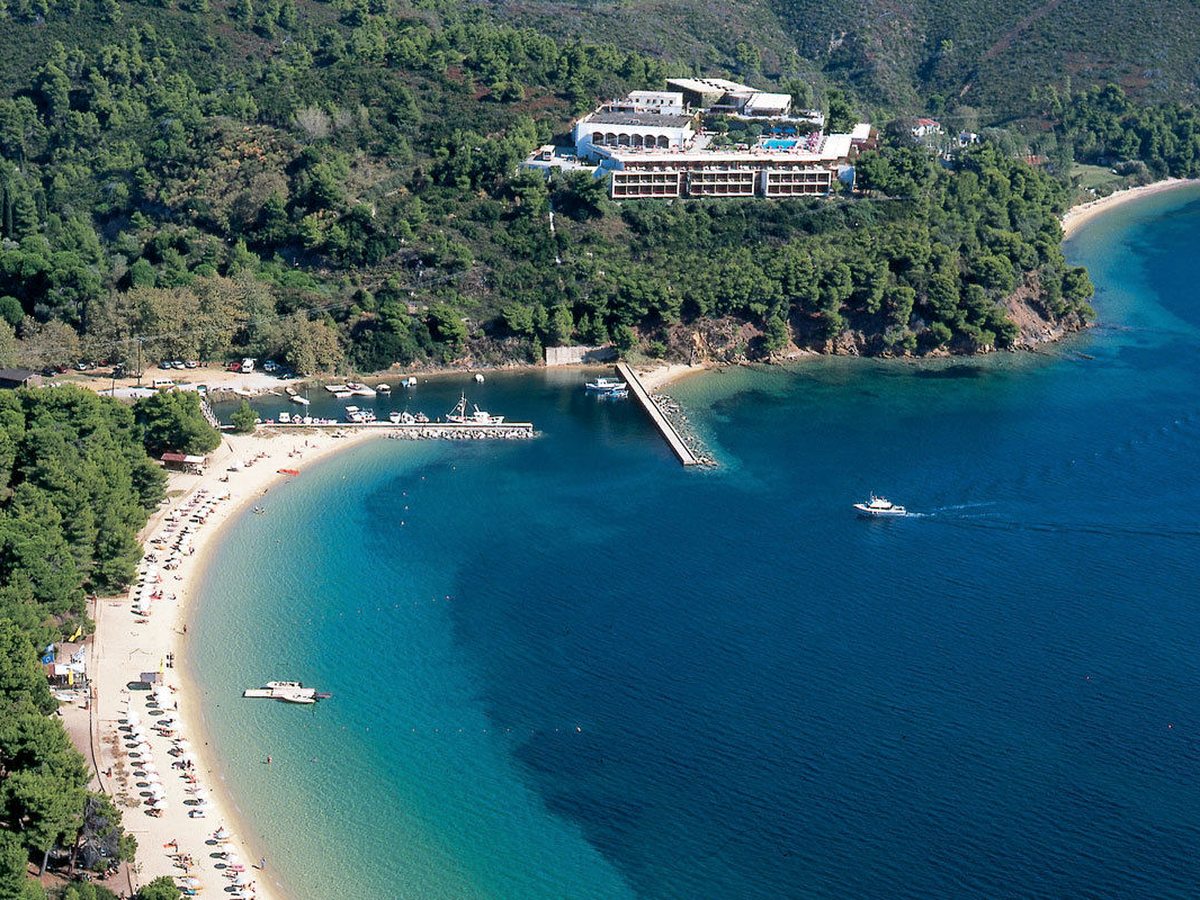 An aerial view of the long white hotel buildings on a pine-covered headland, above a curving sandy beach, a stone jetty and boats moored in a deep blue bay.